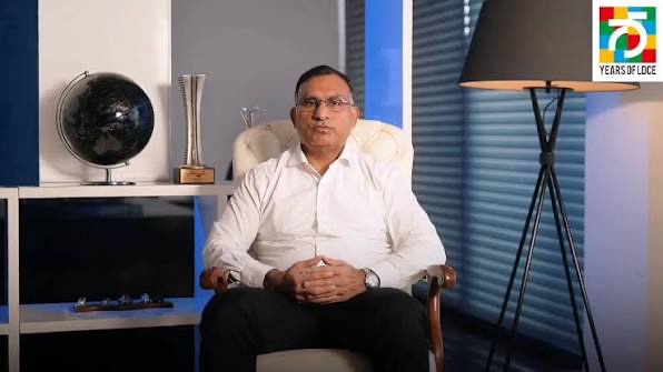 Dhiren Vora seated in a cream armchair in a studio with a globe and trophy on shelves behind him