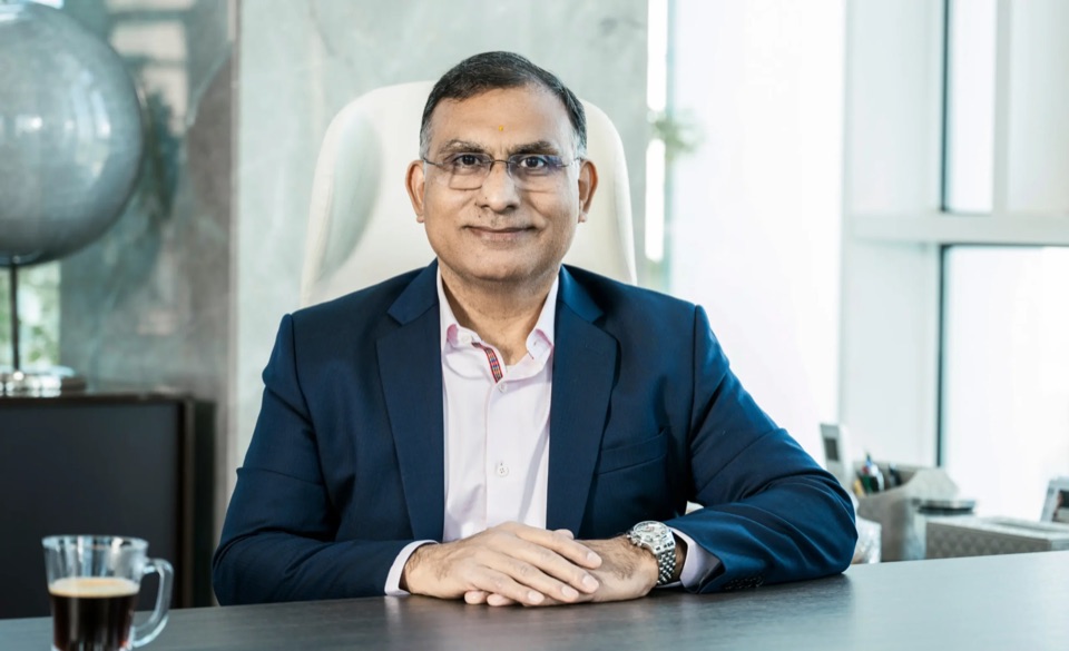 Dhiren Vora seated at a desk with hands folded, a globe and window light behind him